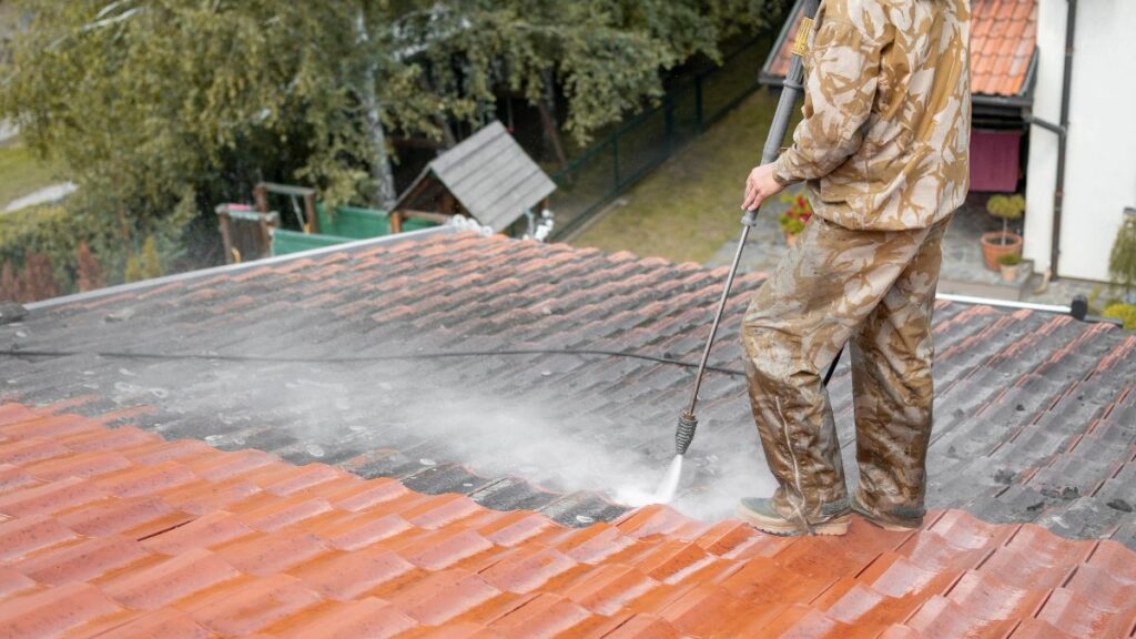 Dakwerker draagt beschermende kledij en reinigt vuile dakpannen met een hogedrukspuit 