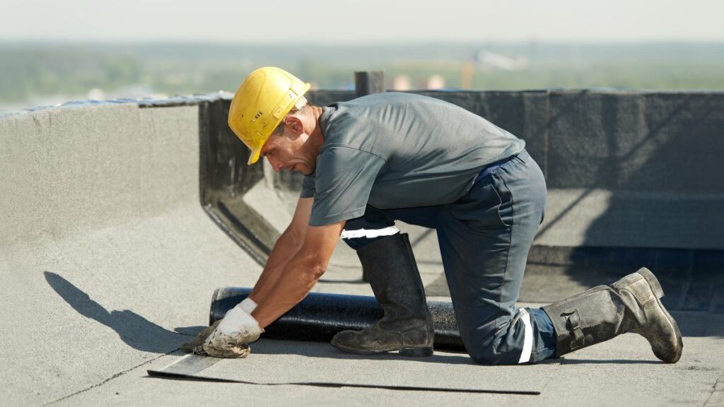 Dakspecialist met gele helm en witte hadnschoenen plaatst een rol bitumen op een plat dak 