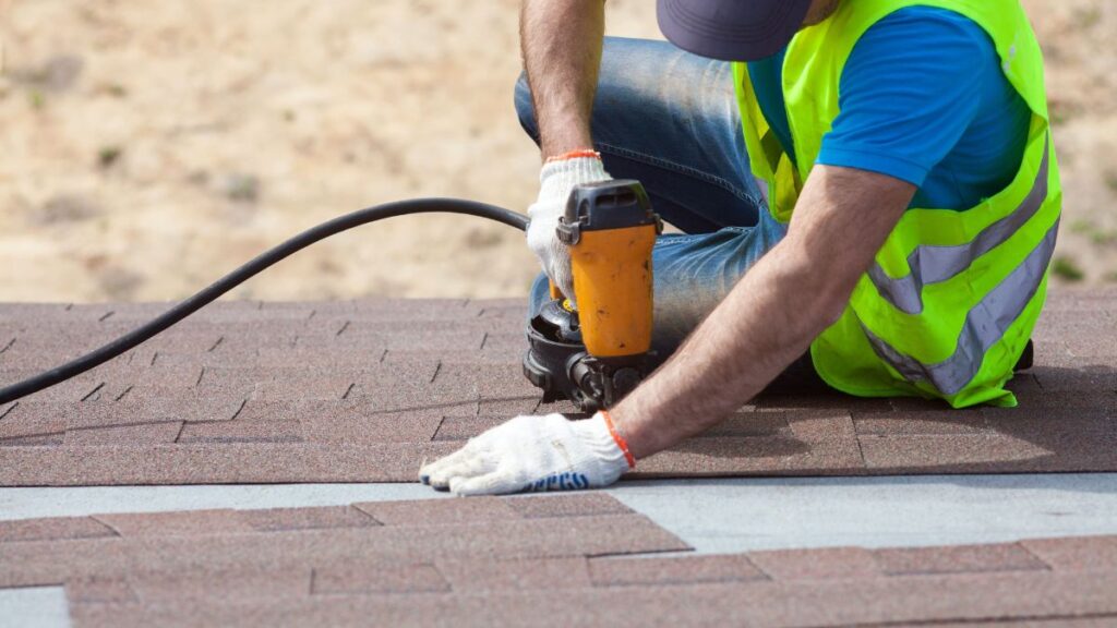 Erkende dakwerker met witte handschoenen, geel fluohesje en pet monteert shingles met een machine op een schuin dak 